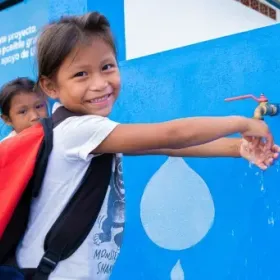Smiling schoolgirl with a backpack washes her hands at an outdoor tap, while another child watches beside a bright blue water mural.