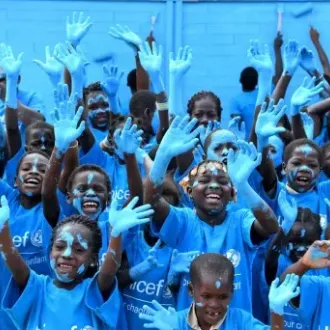 Joyful children covered in blue paint and wearing UNICEF shirts raise their hands before a blue wall, creating an energetic, celebratory scene.