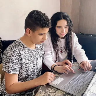Two teenaged youth sit beside each other at a table and work on a shared laptop computer.