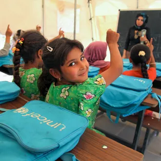 Girl raising her hand in a classroom tent, with UNICEF backpacks on the desks.