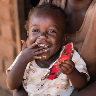 Toddler smiling while eating a therapeutic food packet, held by mother.