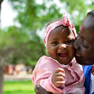 Smiling mother in a blue shirt kisses her baby daughter’s cheek in a park; the baby wears pink and a bow in this joyful, loving scene.