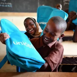 Smiling African schoolchildren hold bright blue UNICEF backpacks in a classroom, conveying joy, hope, and the impact of education aid.