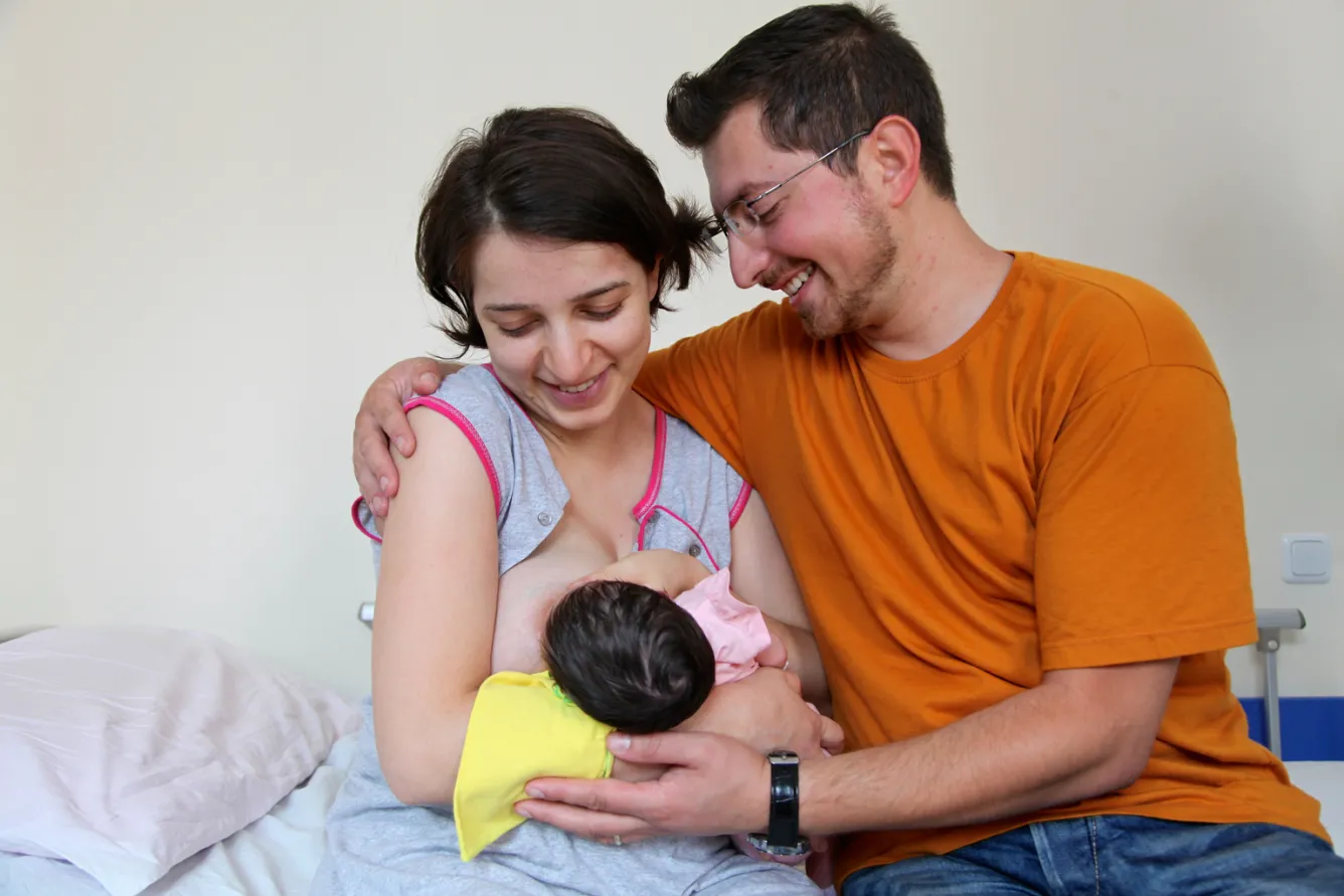 A woman breastfeeds her baby while her husband looks on.