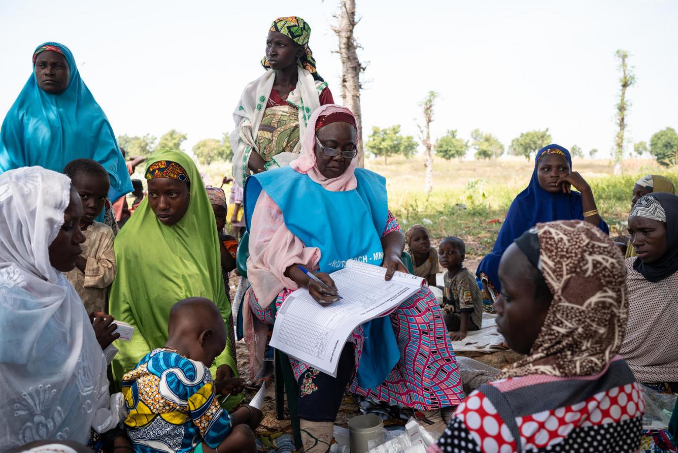 A Nigerian woman wearing a bright blue UNICEF vest sits on a chair among a group of women and children.