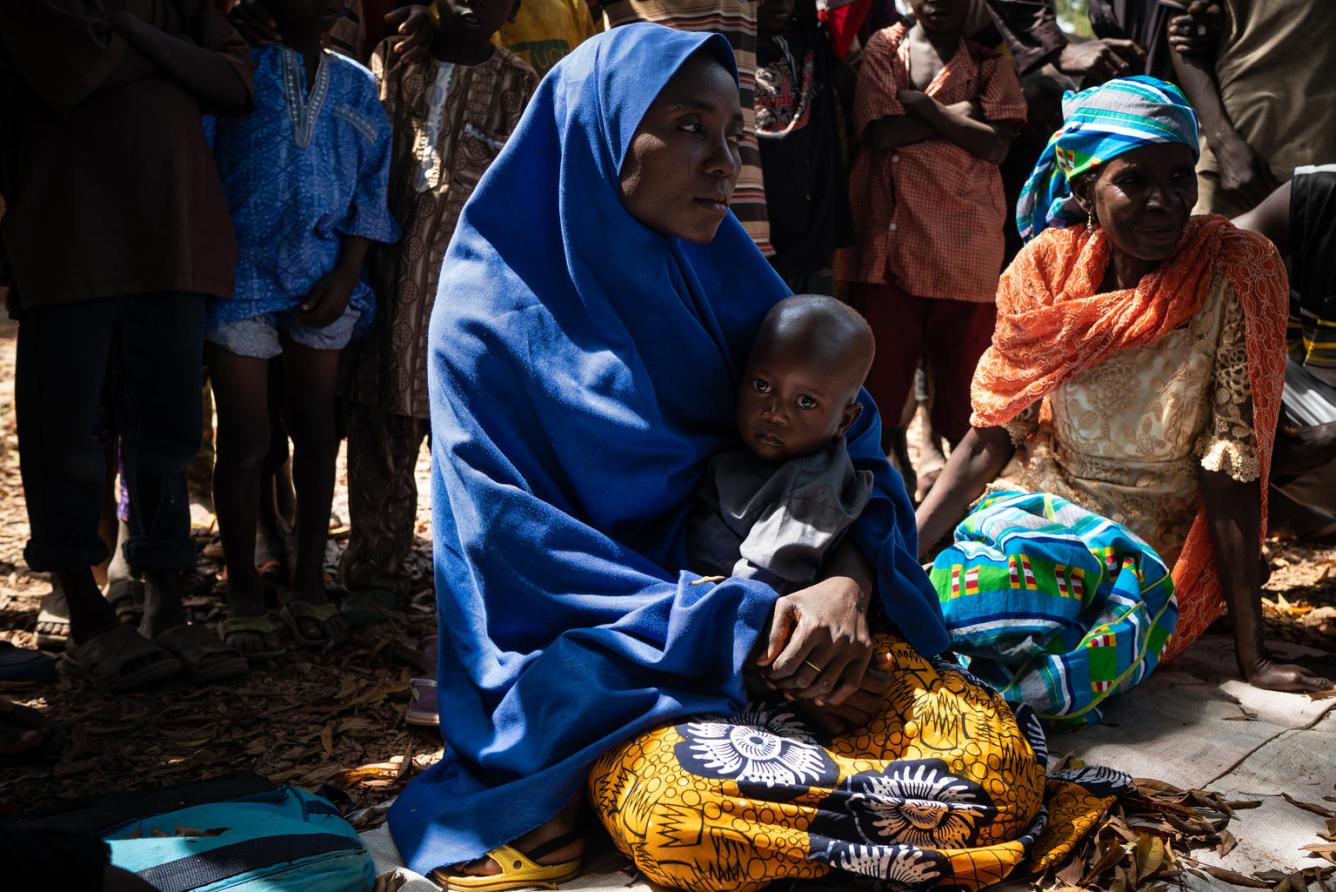 A Nigerian woman in blue head covering sits with a child in lap among a group of women, in Kadura settlement.