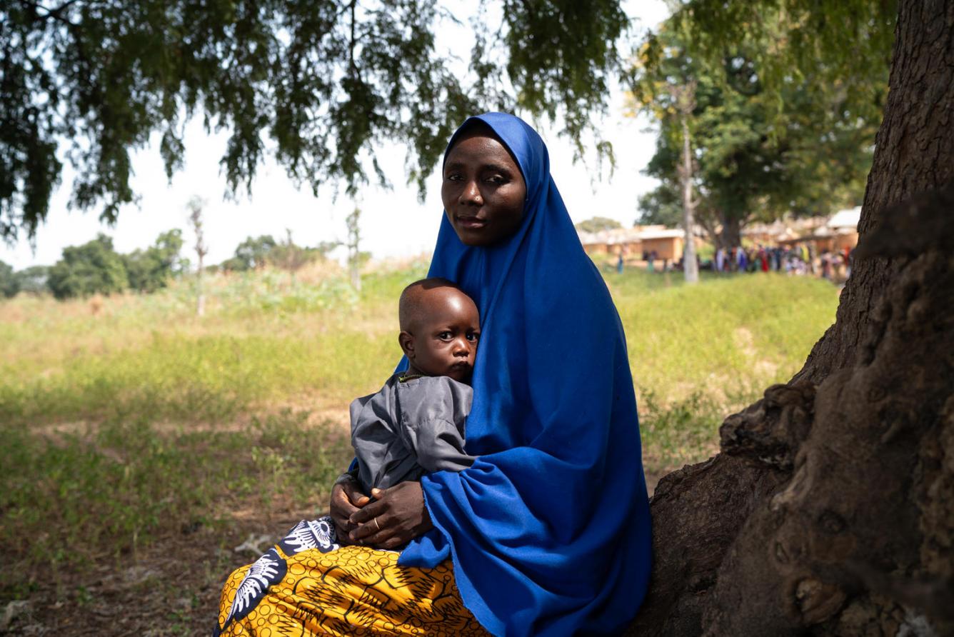 A Nigerian woman in blue head covering sits beside a tree with a child in lap in Kadura settlement.