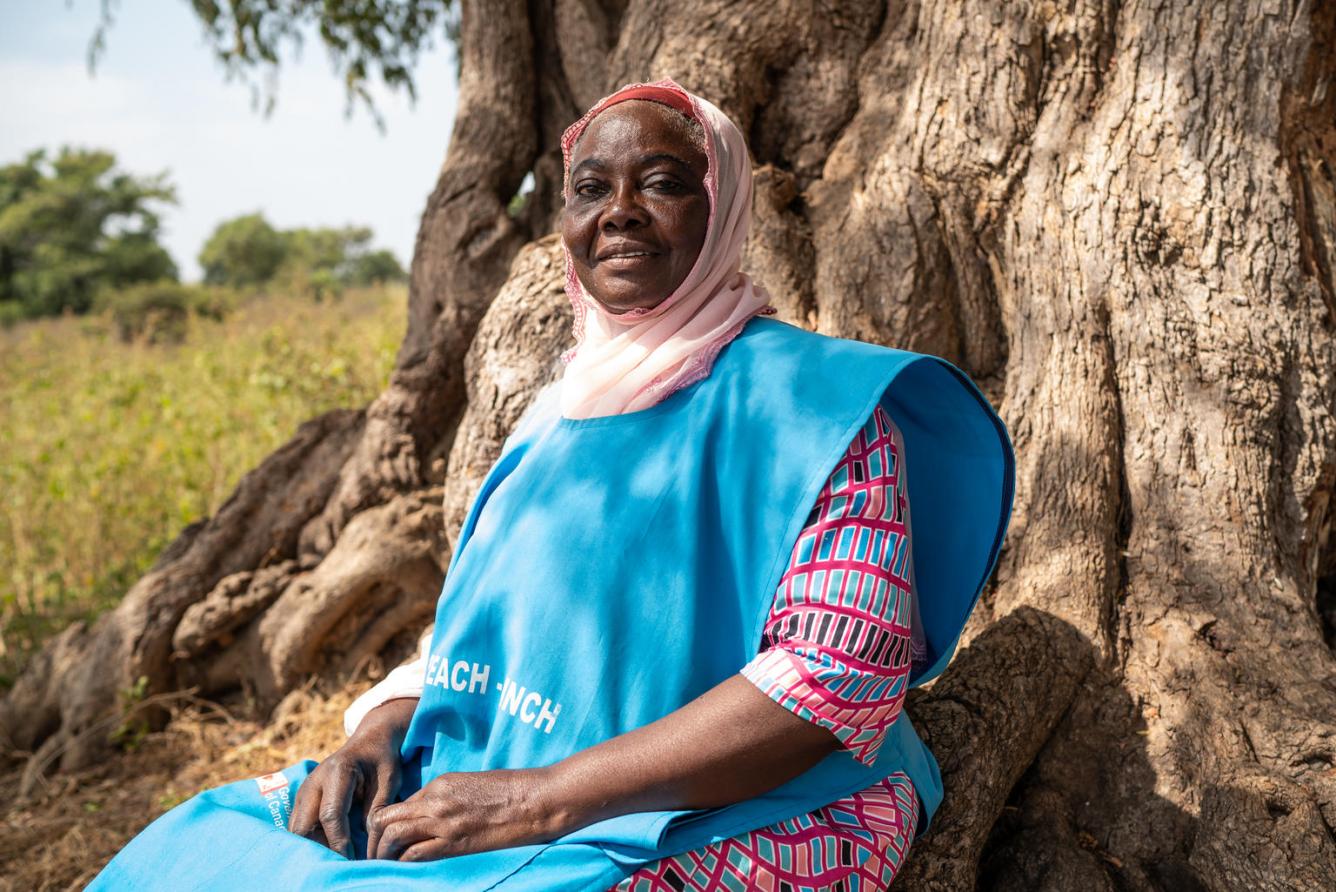 A Nigerian woman sits beside a tree trunk for a portrait in a bright blue UNICEF vest.