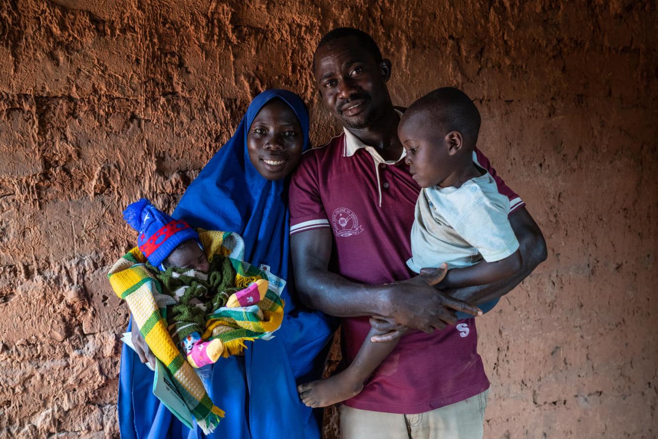 A Nigerian couple with two children pose for a family portrait against a red kuccha wall.
