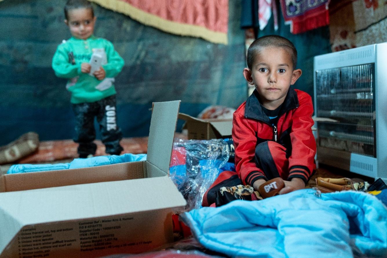 4-year old boy in red jacket looks into the camera, while he opens his new winter clothing kit that he received from UNICEF.