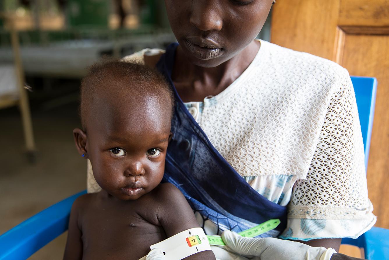 A young child is held by her mother as she gets her arm checked with a malnutrition armband.