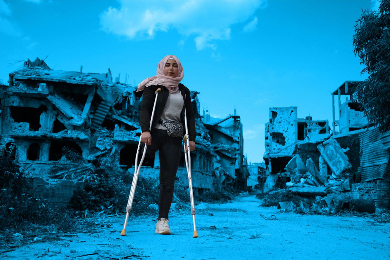 Nour stands using crutches in front of demolished buildings.