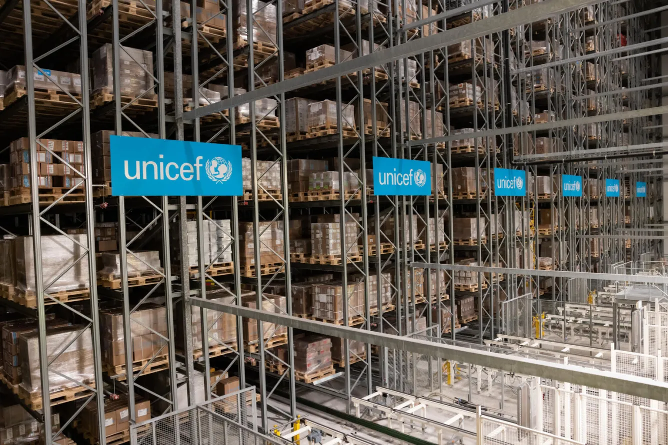 Entrepôt avec logos unicef bleu ciel et chariots empilés jusqu'au plafond avec des boîtes de dons.