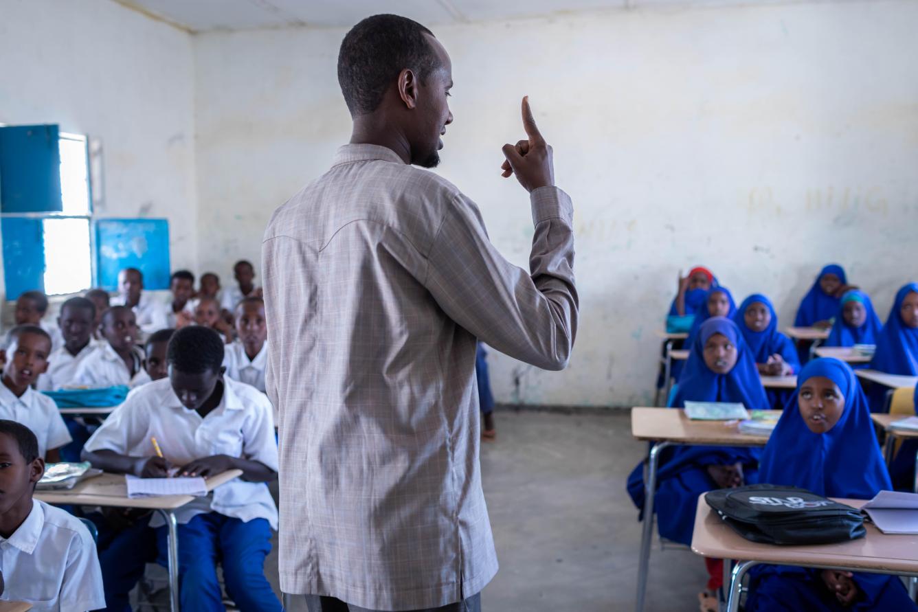 A teacher stands in front of a class, he is holding his finger up, his back is to the camera. In front of him girl students sit on desks on the right. On the left boys sit on desks.