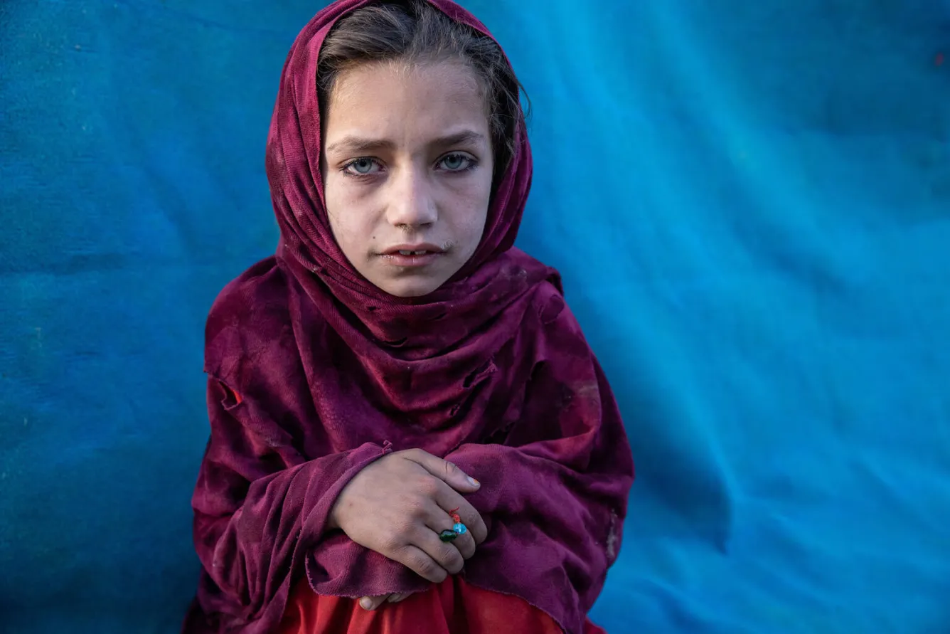 A girl looks directly into a camera, a magenta scarf is around her, her hands are folded over her knees.