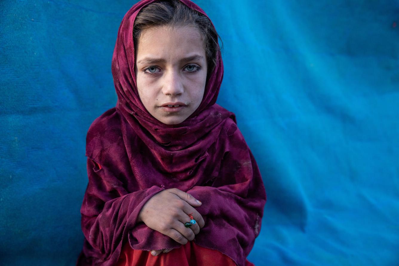A girl looks directly into a camera, a magenta scarf is around her, her hands are folded over her knees.