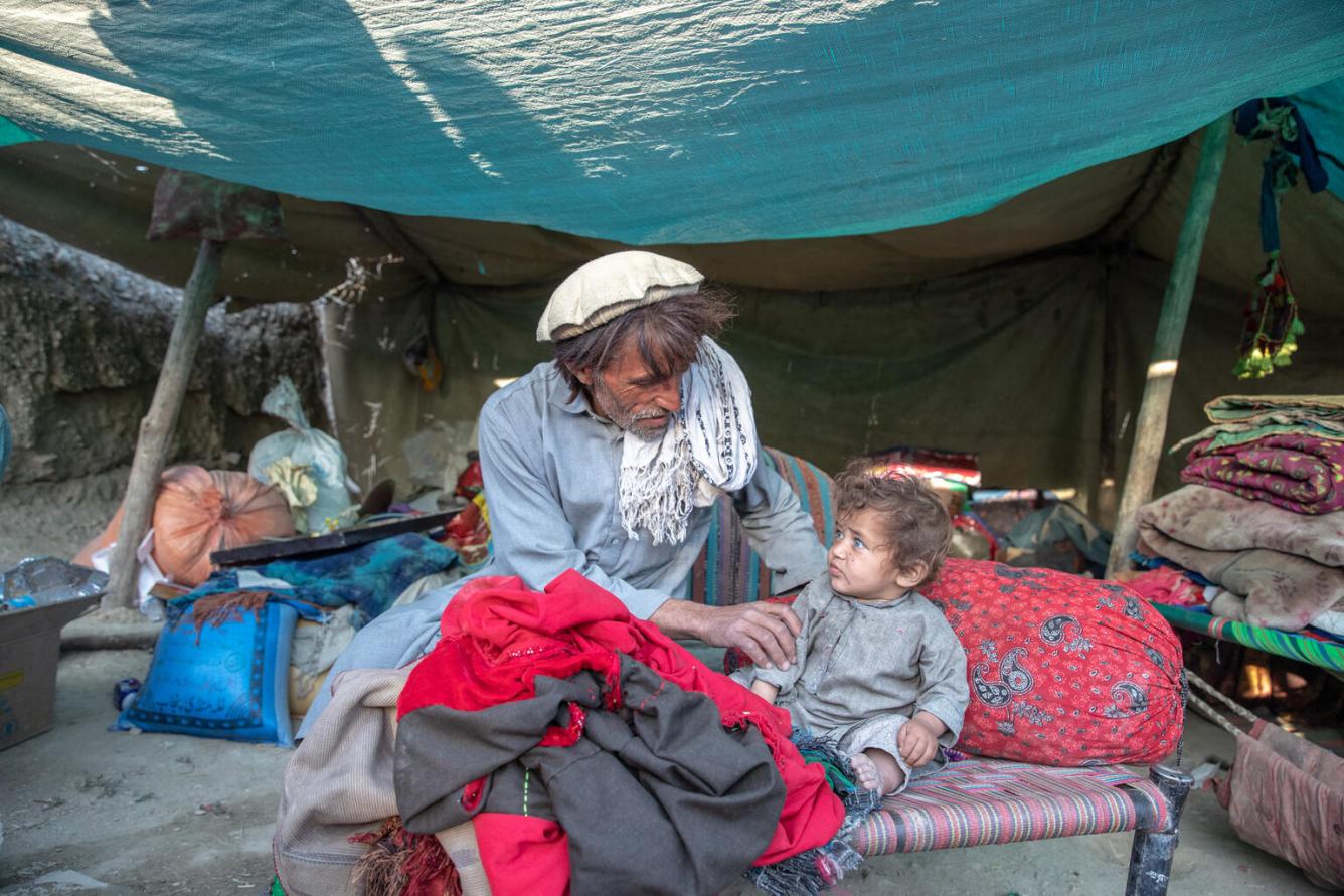 A man sits on a woven bed with an infant next to him. They are looking at each other, behind them is a tent, like a makeshift shelter, with belongings lying in sacks and piles.