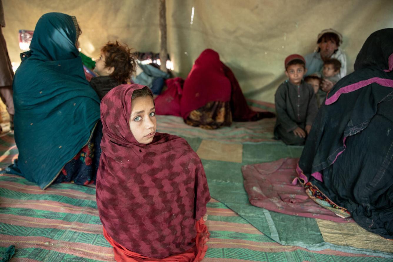 A girl wrapped in a large red scarf sits on the floor of a tent with her head turned back to look at the camera. Around her are other women sitting in a group.