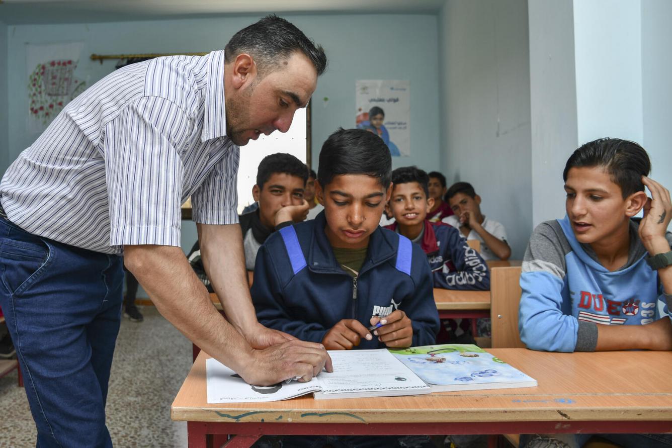A school teacher leans over the desk of two students who have a book open in front of them.