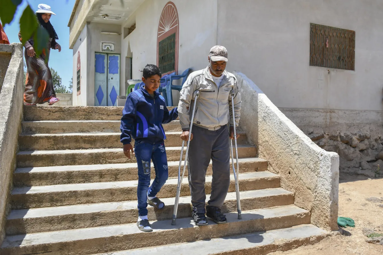 A boy helps a man down a flight of stairs outside. The man has a walking stick in one hand.