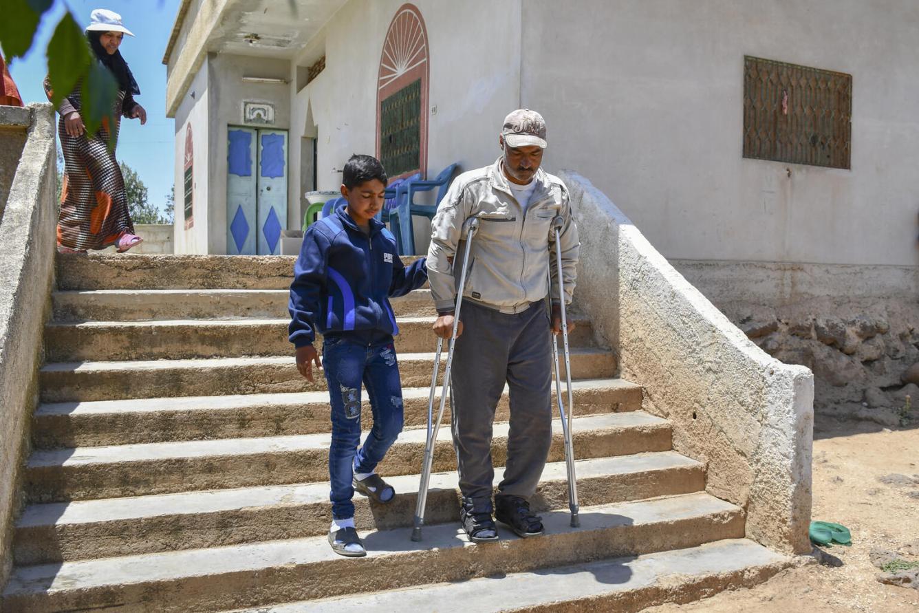 A boy helps a man down a flight of stairs outside. The man has a walking stick in one hand.