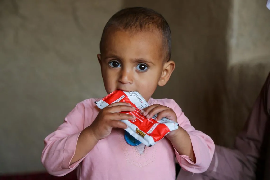 A toddler in pink clothing is holding a red packet of ready-to-eat-therapeutic food. The photo is zoomed in, we can see the toddler from the chest up. He is looking at the camera.