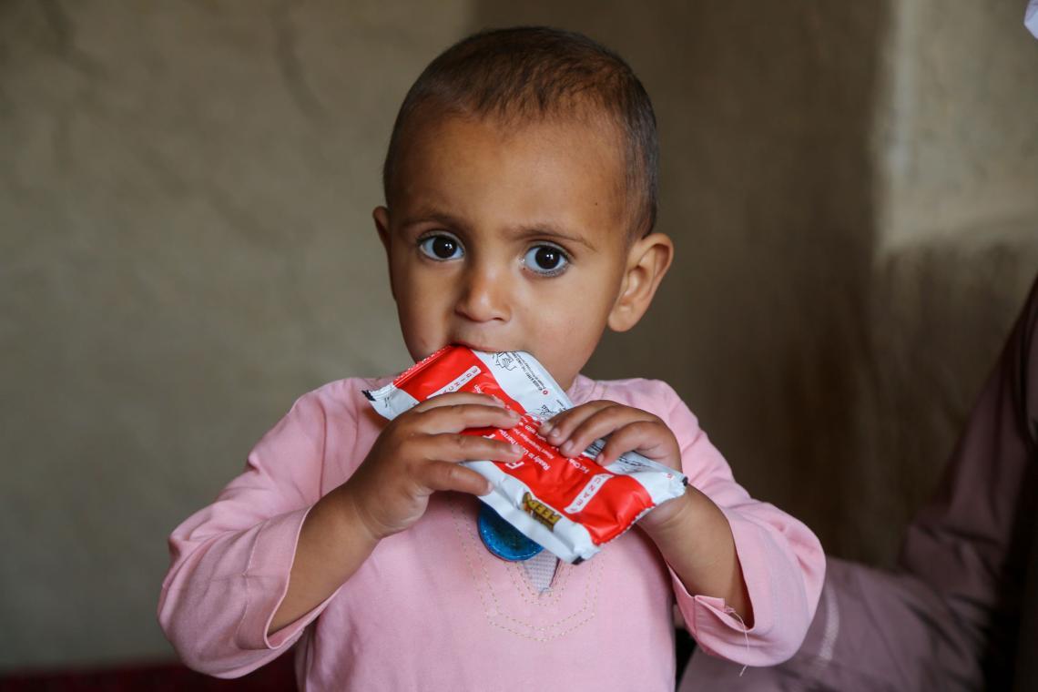 A toddler in pink clothing is holding a red packet of ready-to-eat-therapeutic food. The photo is zoomed in, we can see the toddler from the chest up. He is looking at the camera.