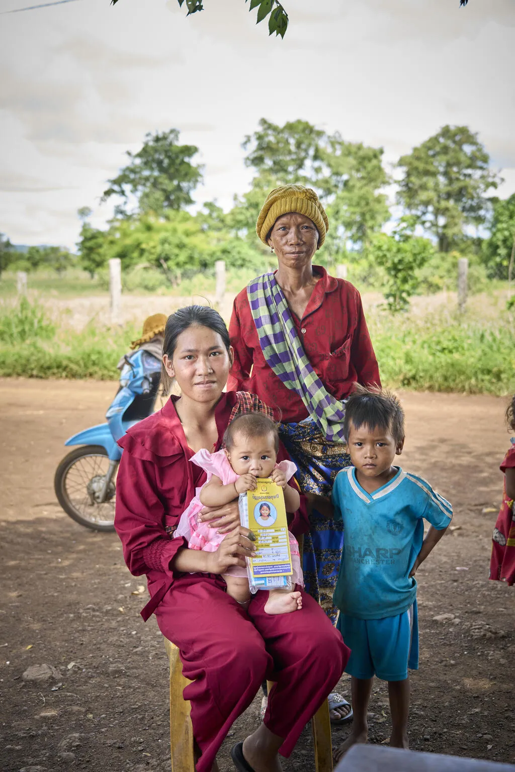 A young woman is seated outdoors, holding an infant in her arms. Behind her an elderly woman stands and in front of them is a boy in blue clothing also standing. They are all looking at the camera. The infant in gnawing on a vaccination card.