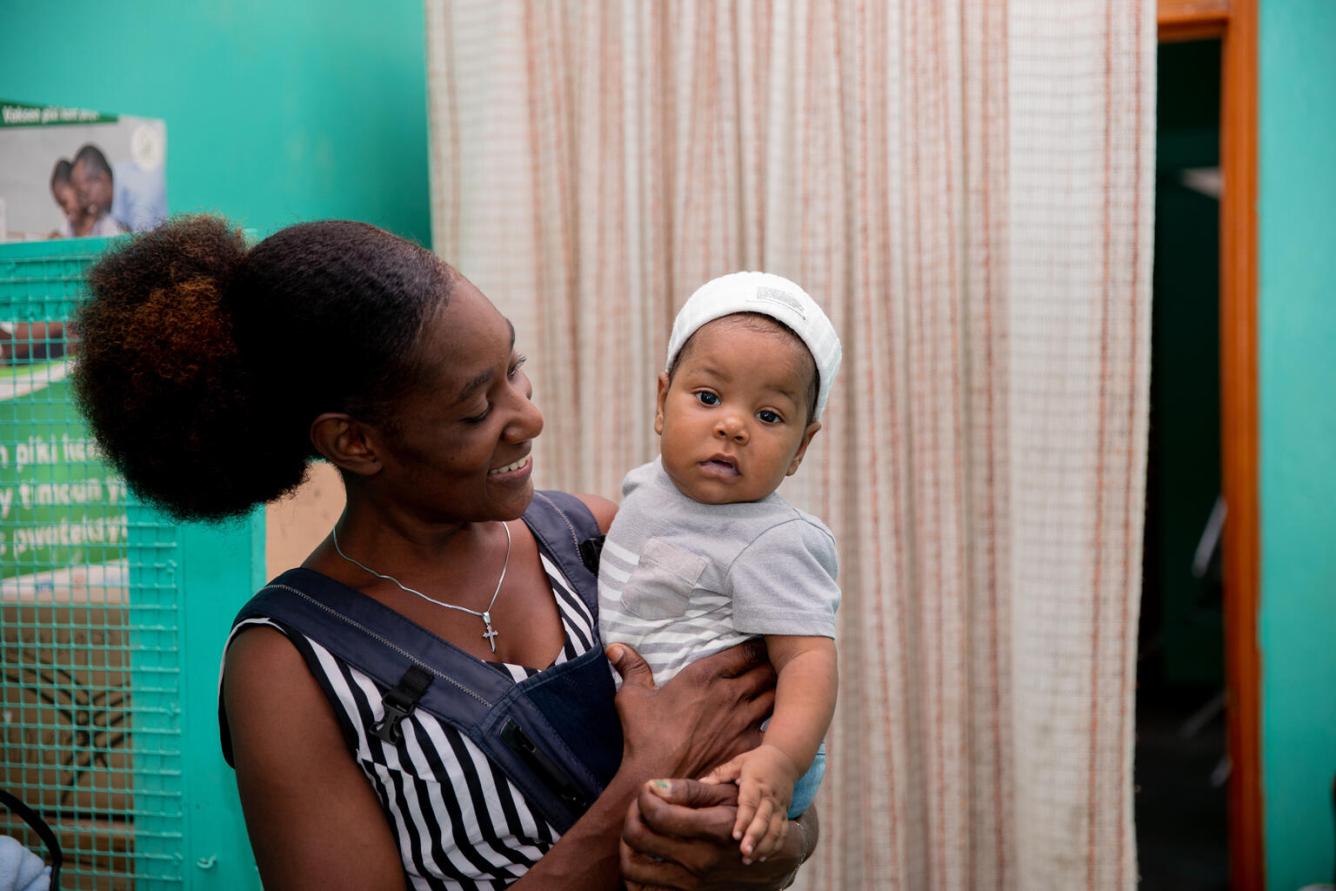 A woman holds an infant and is smiling. The infant is looking at the camera while the woman is looking at the child. They are in a clinic-looking setting.