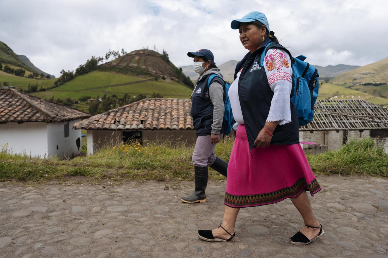 Two women walk on a cobbled path, with a mountainous terrain behind them. The woman in the foreground is wearing a blue UNICEF cap.