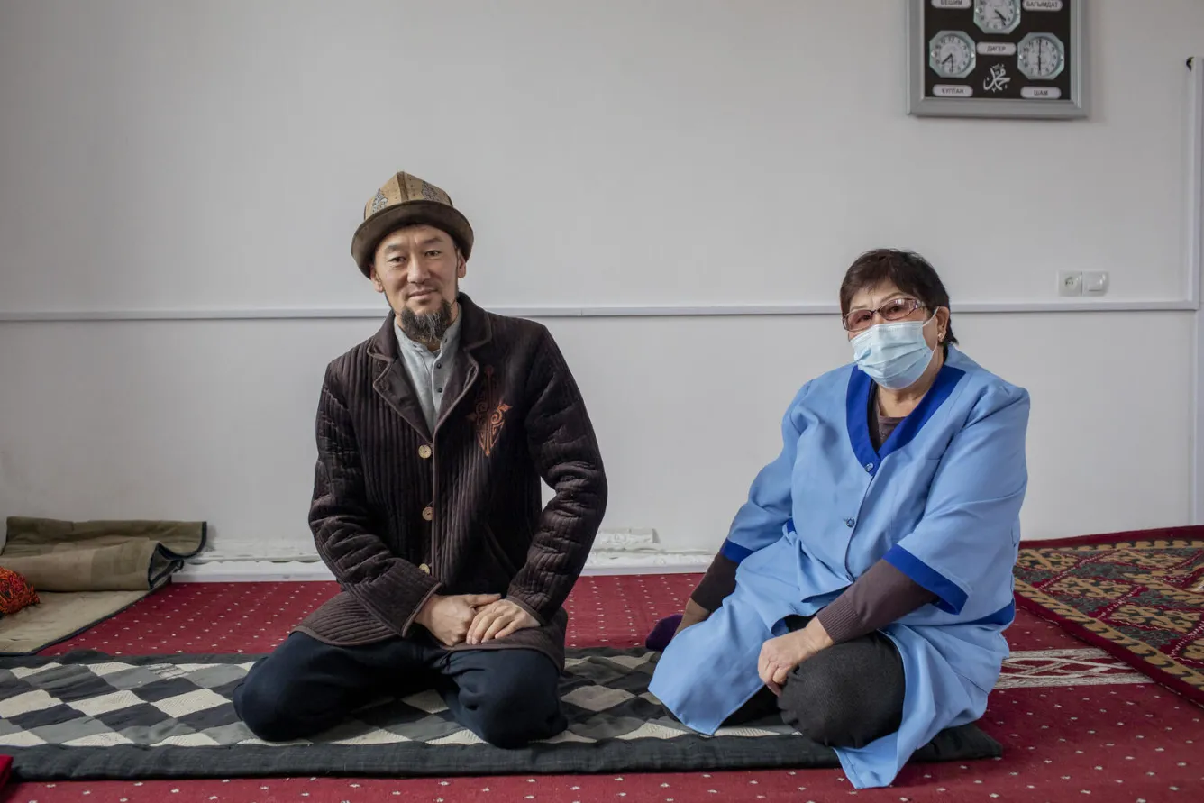 A man in traditional Kyrgyz clothng sits in a mosque with a woman next to him who is wearing a blue nurse's uniform and a mask. They are both seated on a carpeted floor looking straight at the camera.