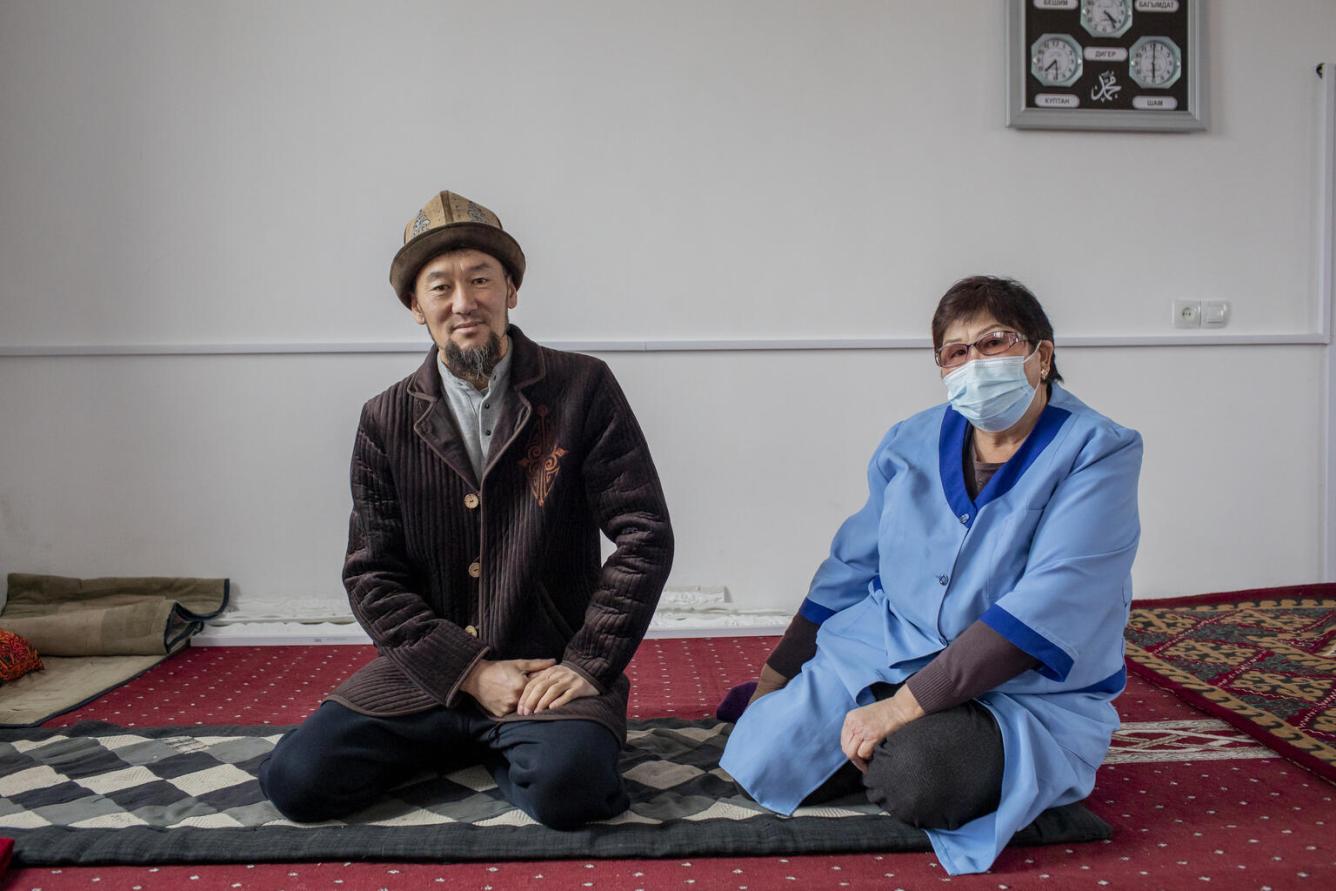 A man in traditional Kyrgyz clothng sits in a mosque with a woman next to him who is wearing a blue nurse's uniform and a mask. They are both seated on a carpeted floor looking straight at the camera.