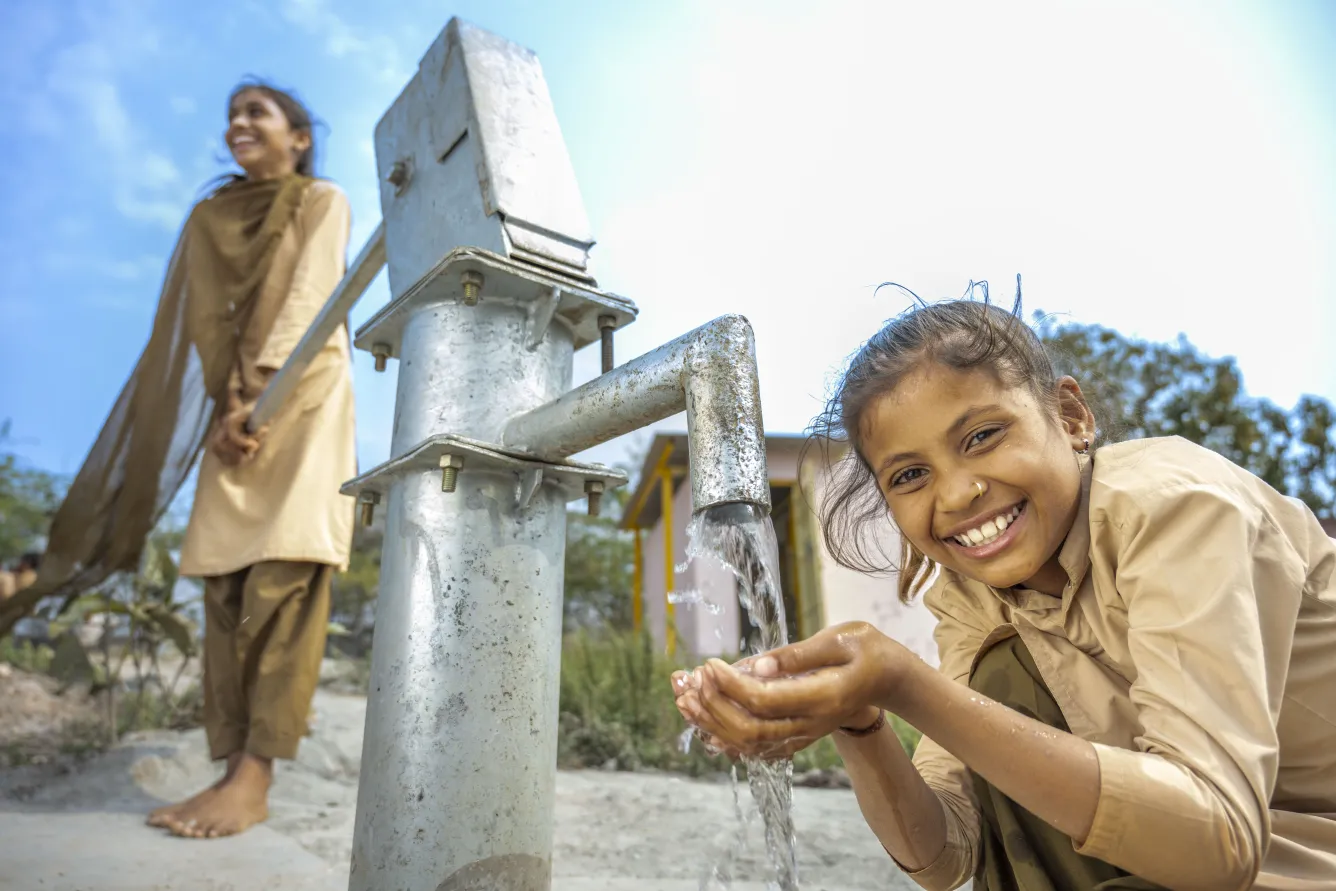 In India, a young girl smiles while holding her hands under the tap of a water pump while another girl works the pump handle in the background, smiling.