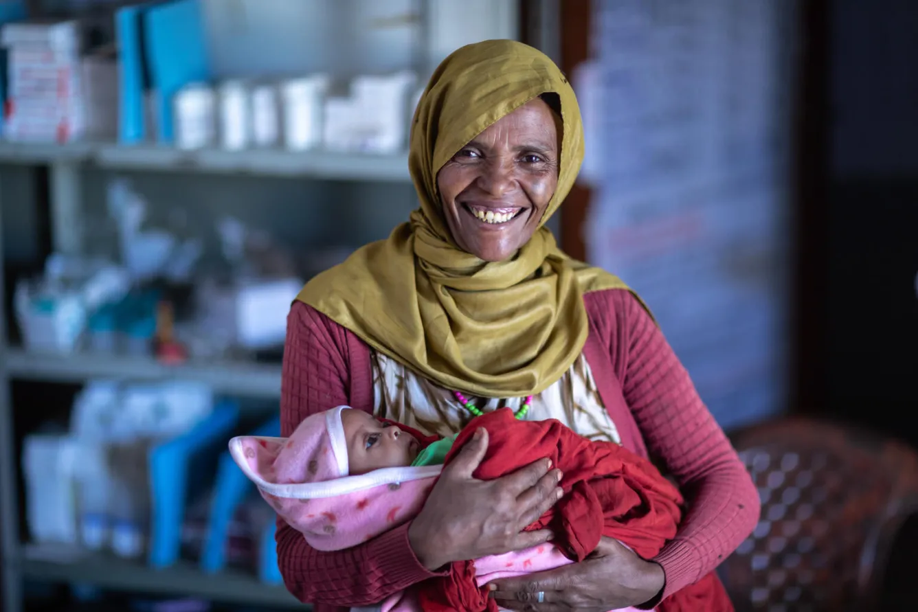 A woman looks at the camera smiling. She is holding a baby in a pink blanket who is sleeping.