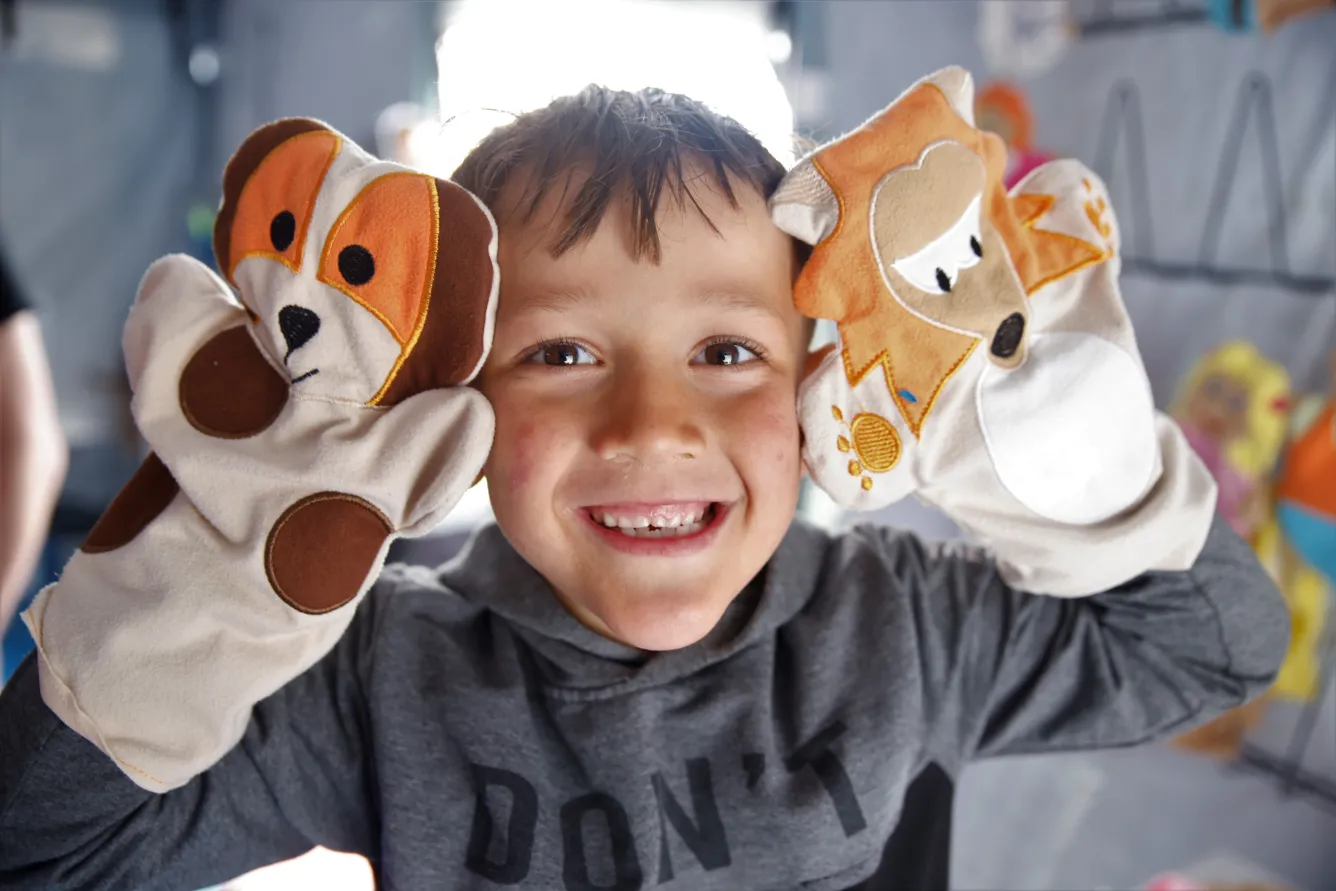 A boy looks at the camera and smiles while waving two hand puppets.