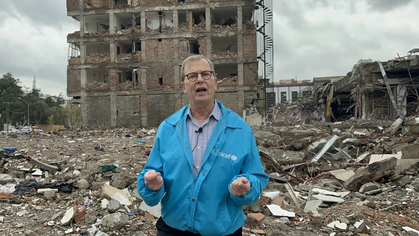 A man wearing a blue UNICEF jacket stands in front of a rubble, behind him the ruins of a building.