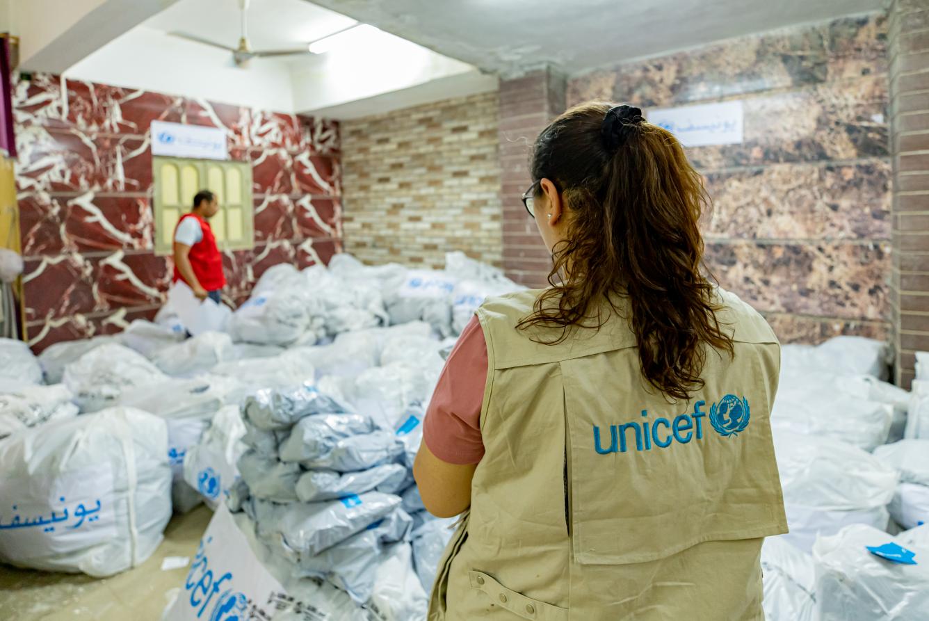 A woman looks at a room full of bags with UNICEF logos on them. Her back is turned to the camera and she is wearing a khaki vest with the UNICEF logo written at the back..
