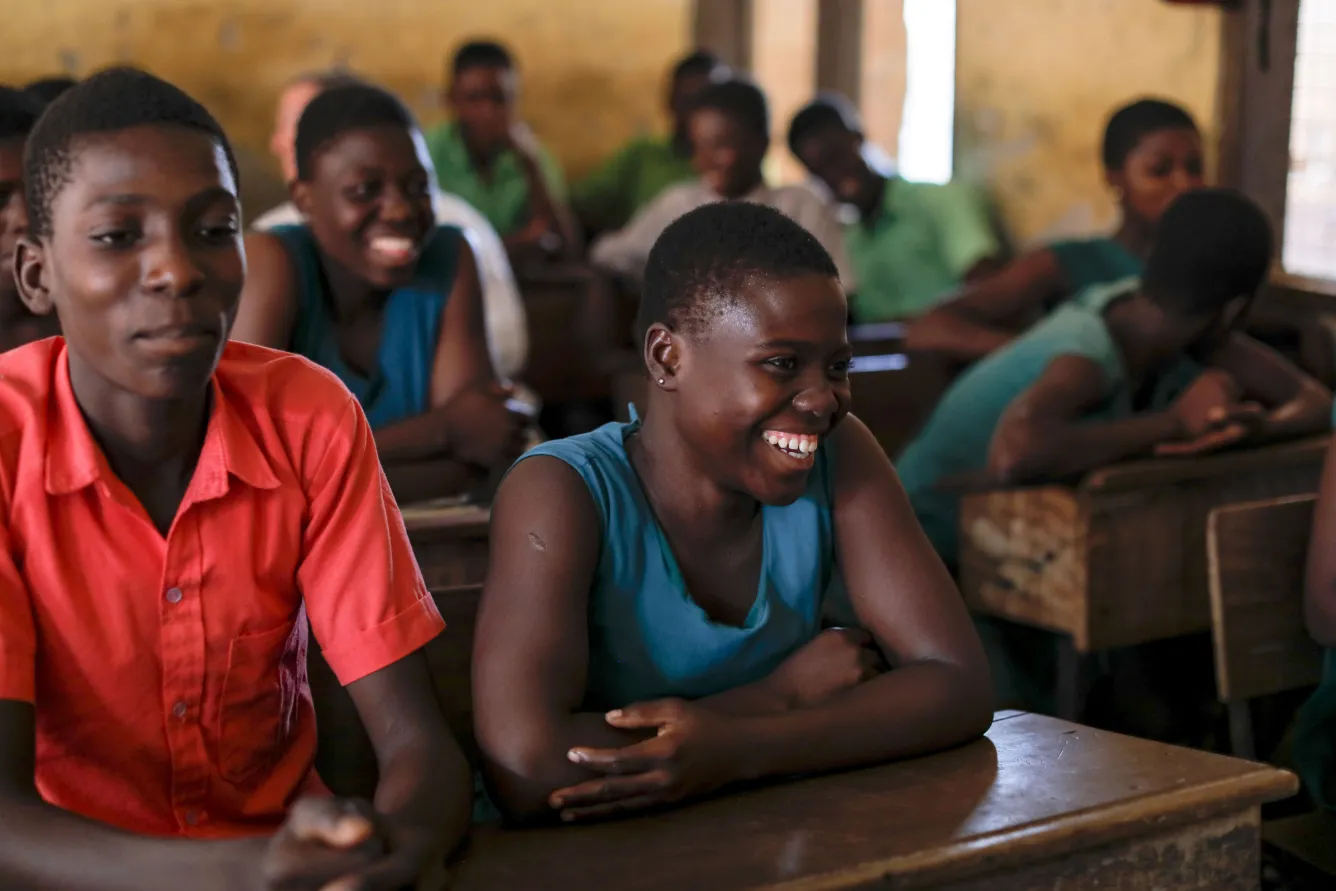 A classroom of girls in Ghana. IN the foreground two adolescent girls sit, one of the left is laughing, the one on the right is looking ahead.