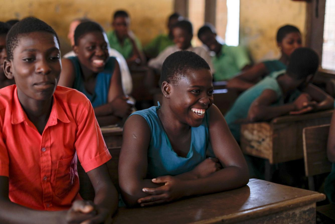 A classroom of girls in Ghana. IN the foreground two adolescent girls sit, one of the left is laughing, the one on the right is looking ahead.