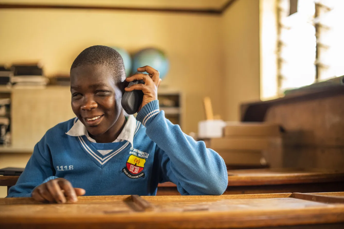 A girl inside a classroom wearing a blue uniform sweater holds up an accessibility reader to her ear as she smiles in listens in Uganda.