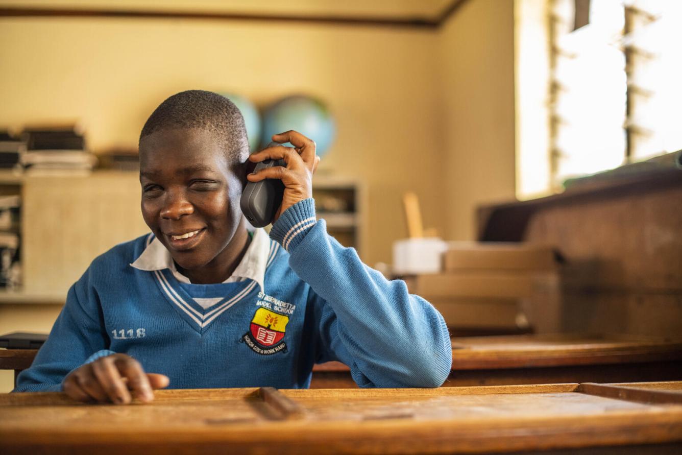 A girl inside a classroom wearing a blue uniform sweater holds up an accessibility reader to her ear as she smiles in listens in Uganda.