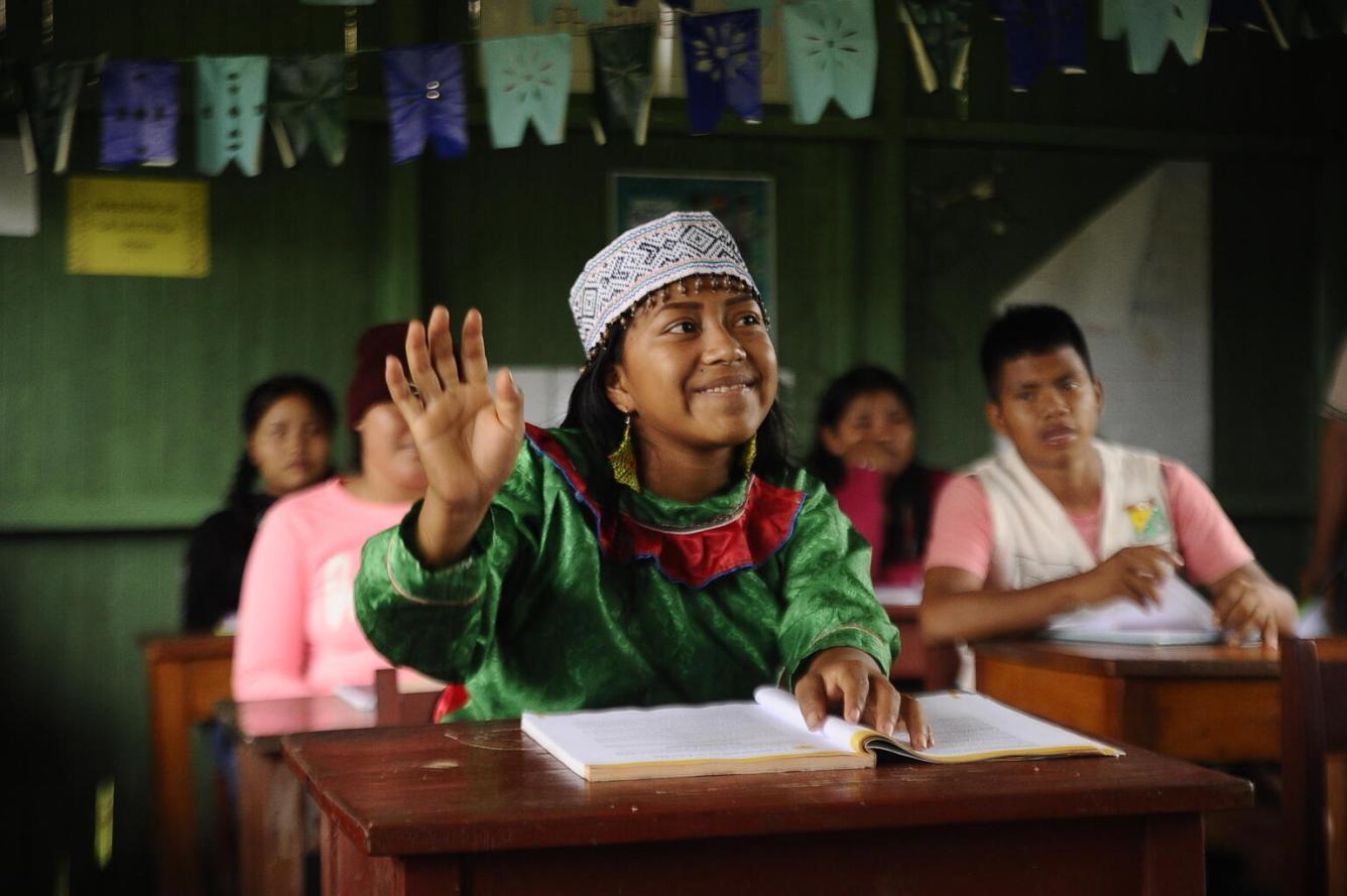 A girl sits at a desk in a classroom with others behind her. She is raising her hand and smiling while wearing traditional Peruvian headband.