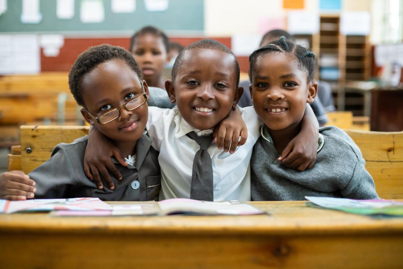 Three boys in grey and white school uniforms sitting at a desk in a classroom with their hands fling around each others necks smile for the camera. In South Africa.