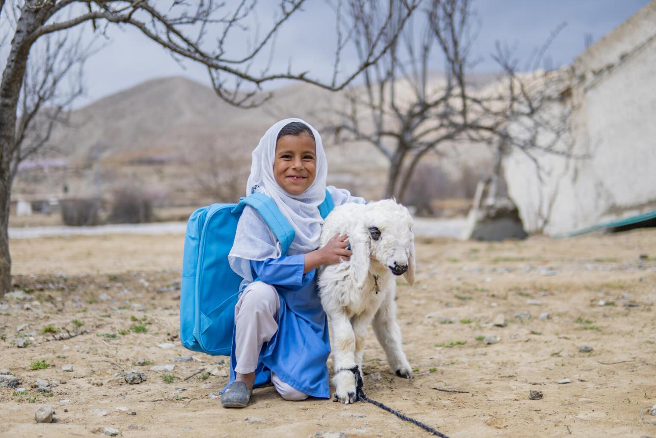 A girl in a blue uniform and UNICEF bag pack crouches next to a lamb in an outdoor setting, looking at the camera and smiling. In Balochinstan, Pakistan.