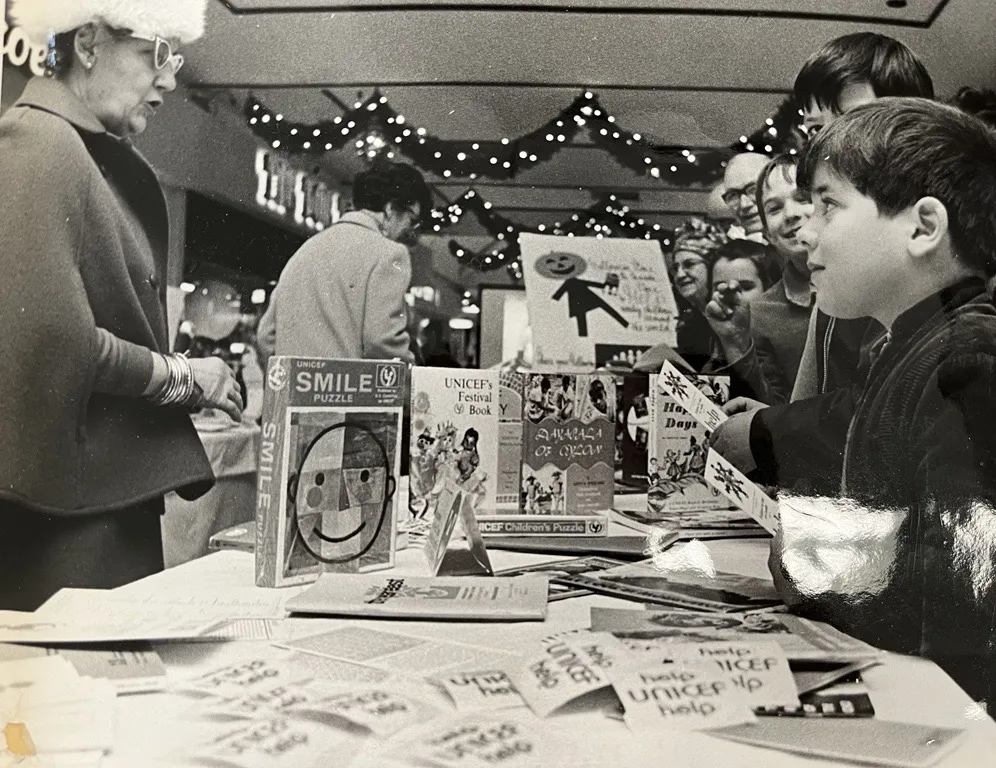 Photo en noir et blanc de deux enfants regardant une femme derrière une table qui vend des articles de l’UNICEF.