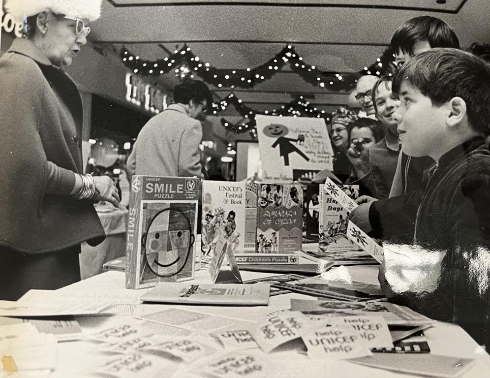 Photo en noir et blanc de deux enfants regardant une femme derrière une table qui vend des articles de l’UNICEF.