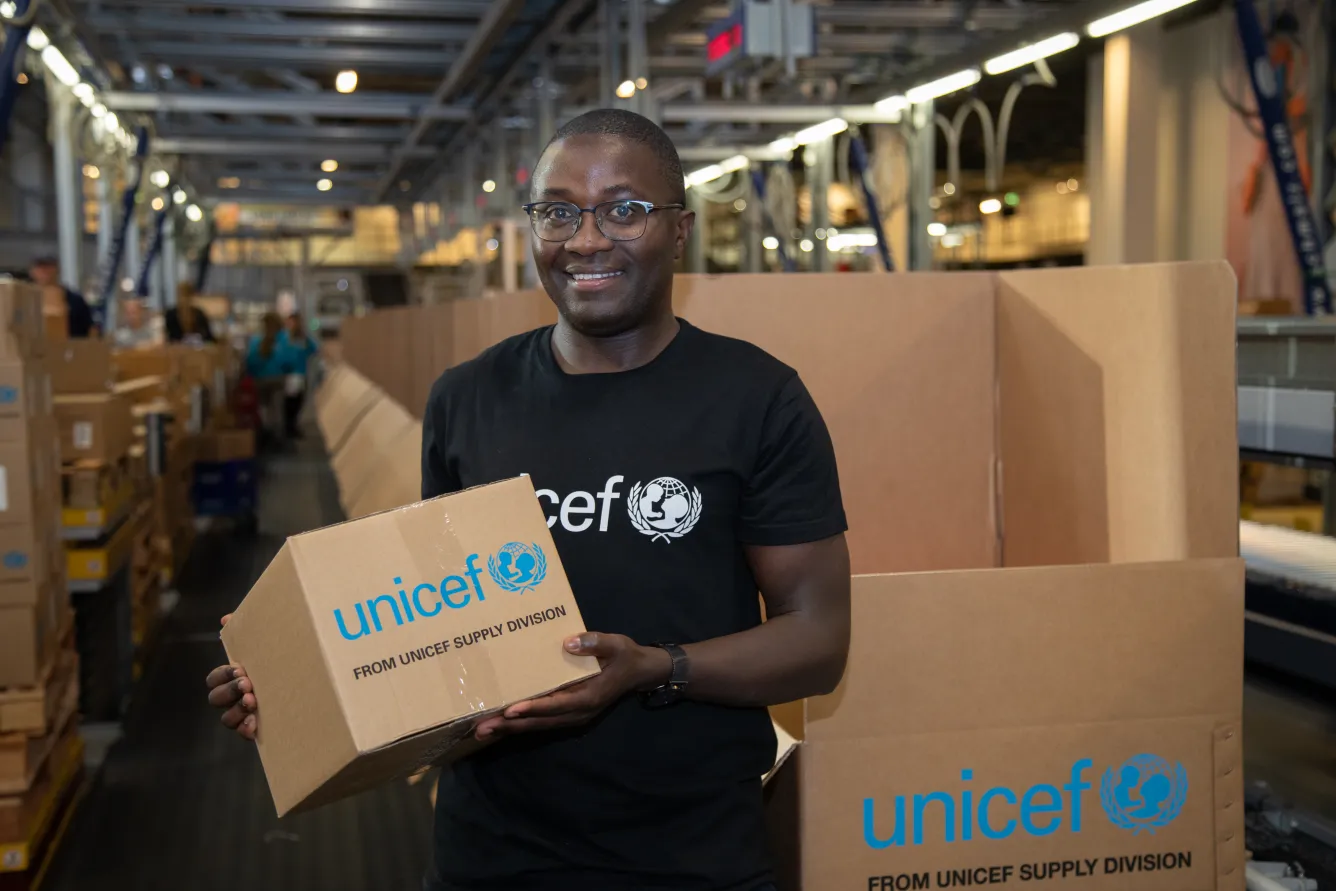 A man holds a cardboard box that says UNICEF on it, behind him more boxes are stacked.