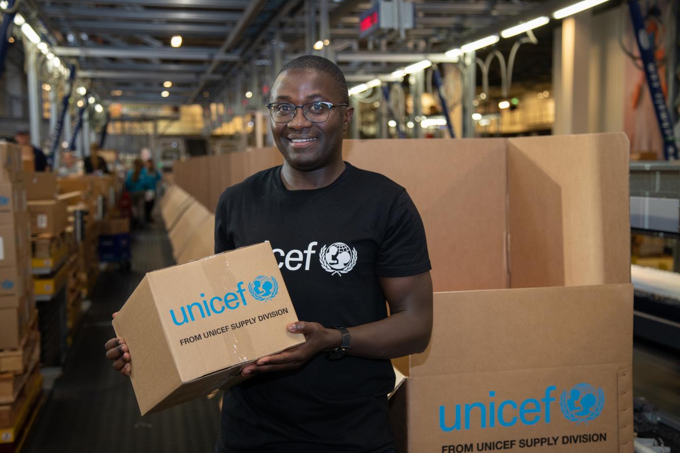 A man holds a cardboard box that says UNICEF on it, behind him more boxes are stacked.
