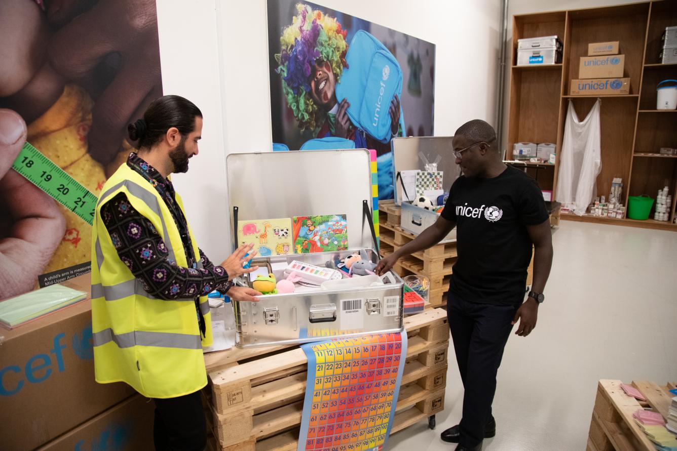 Two men stand besides a metal box that is open and full of school supplies.