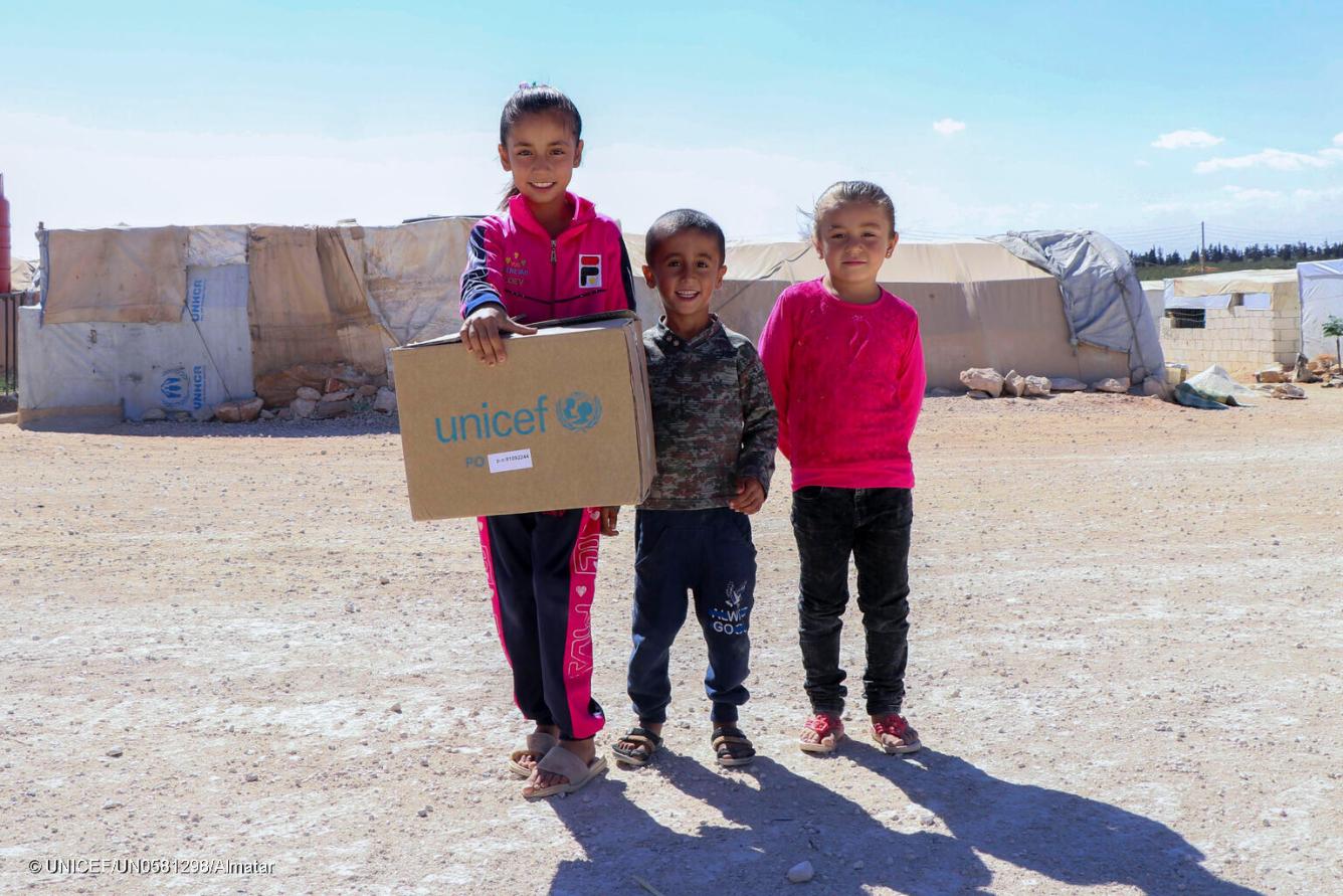 Deux jeunes filles, l’une tenant une boîte de l’UNICEF, et un garçon se tiennent dans un camp de personnes réfugiées à Alep, en Syrie.